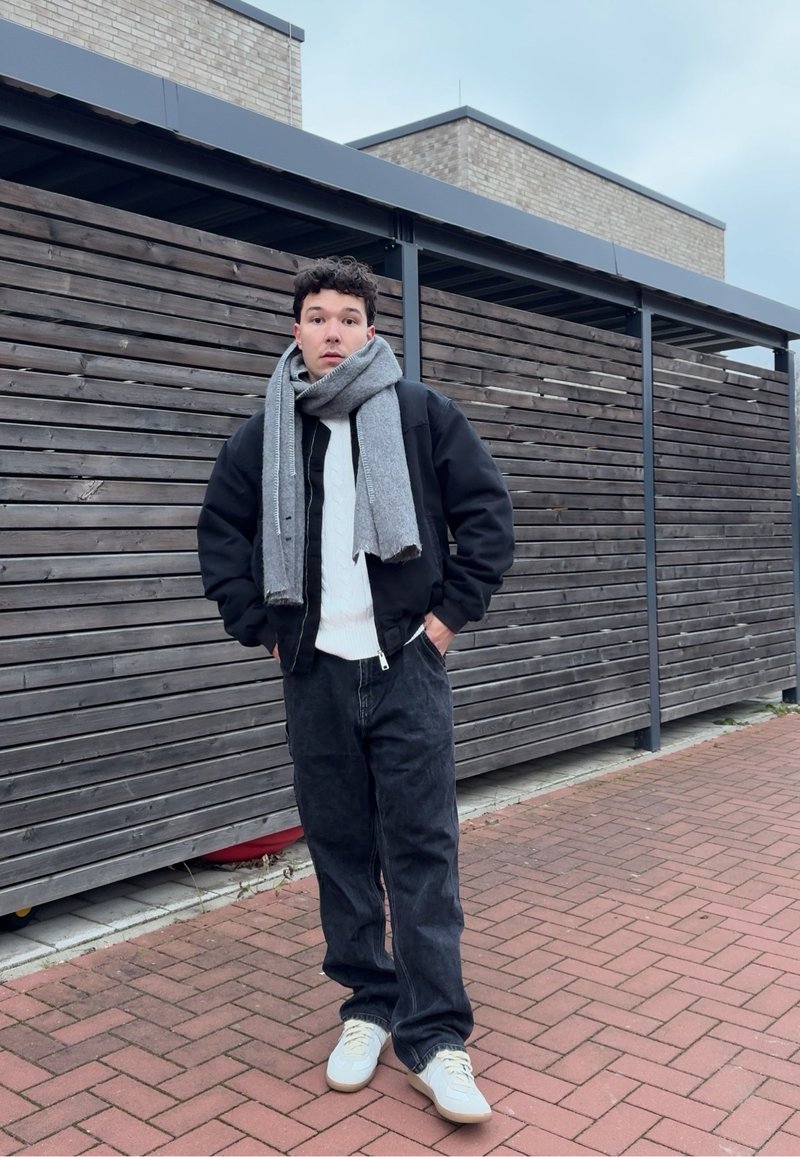 Schwarze Bomberjacke über einem weißen Zopfmuster-Pullover, grauer Schal, dunkle Jeans und beige Turnschuhe, vor einem Holz-Hintergrund und Bürgersteig.