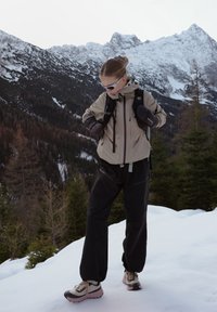 Femme portant une veste beige, un pantalon noir et des lunettes de soleil, ajustant les bretelles de son sac à dos tout en se tenant sur la neige avec une montagne enneigée et des arbres en arrière-plan.