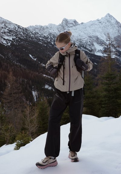 Mujer con chaqueta beige, pantalones negros y gafas de sol ajustándose las correas de la mochila mientras está de pie sobre la nieve, con montañas nevadas y árboles al fondo.