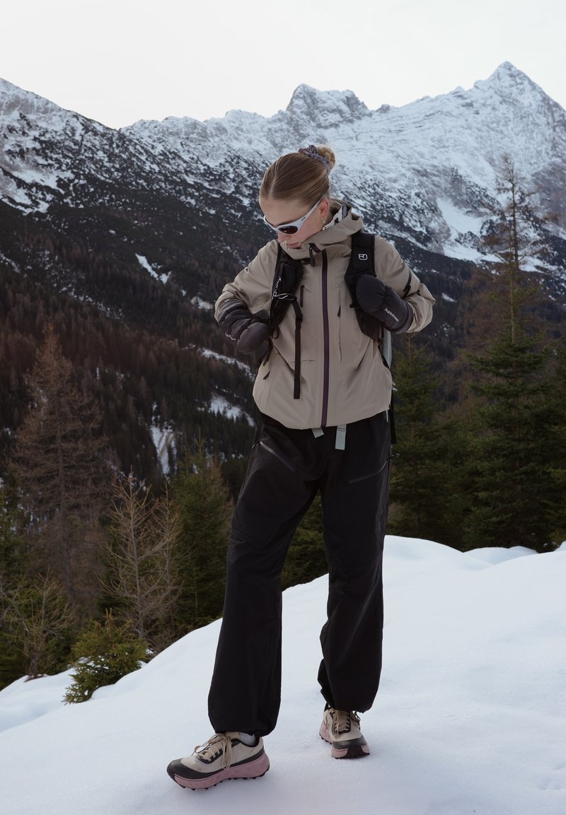 Femme portant une veste beige, un pantalon noir et des lunettes de soleil, ajustant les bretelles de son sac à dos tout en se tenant sur la neige avec une montagne enneigée et des arbres en arrière-plan.