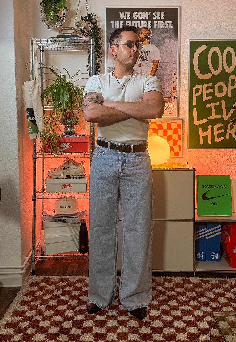 Jean large en denim délavé clair associé à un t-shirt blanc ajusté. Accessoires : ceinture noire et lunettes de soleil. En arrière-plan, des étagères avec des baskets et des décorations.