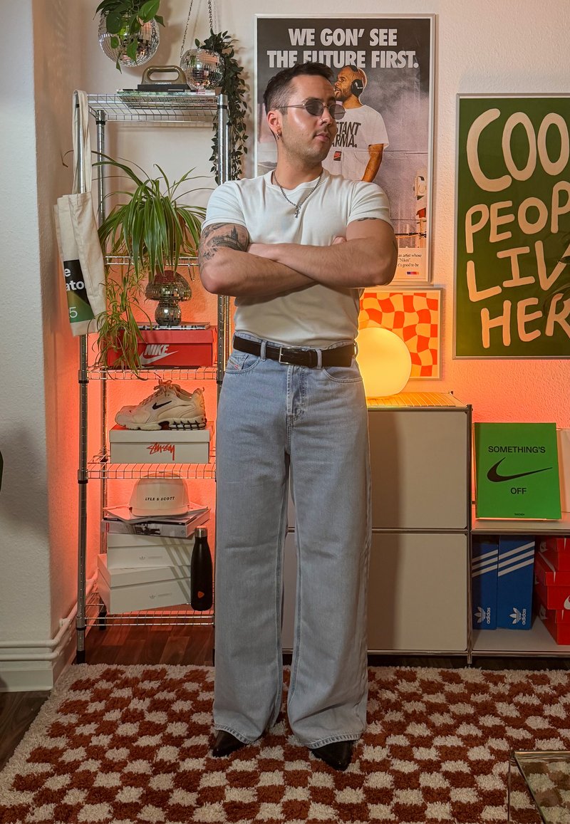 Jean large couleur denim clair associé à un t-shirt blanc ajusté. Accessoires : ceinture noire et lunettes de soleil. En arrière-plan, des étagères avec des baskets et des objets décoratifs.