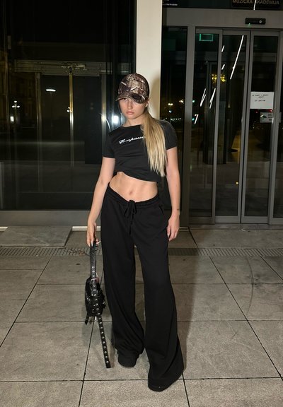 Camiseta negra corta con texto blanco, pantalones negros relajados y una gorra de béisbol en camuflaje. Sosteniendo una bolsa negra de transporte con correas.