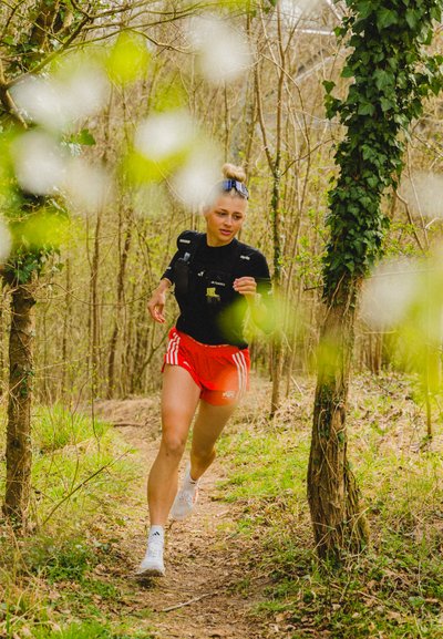 Corredora vestida con una camiseta negra y pantalones cortos rojos con rayas blancas, usando zapatillas blancas, navegando por un sendero boscoso con vegetación verde.