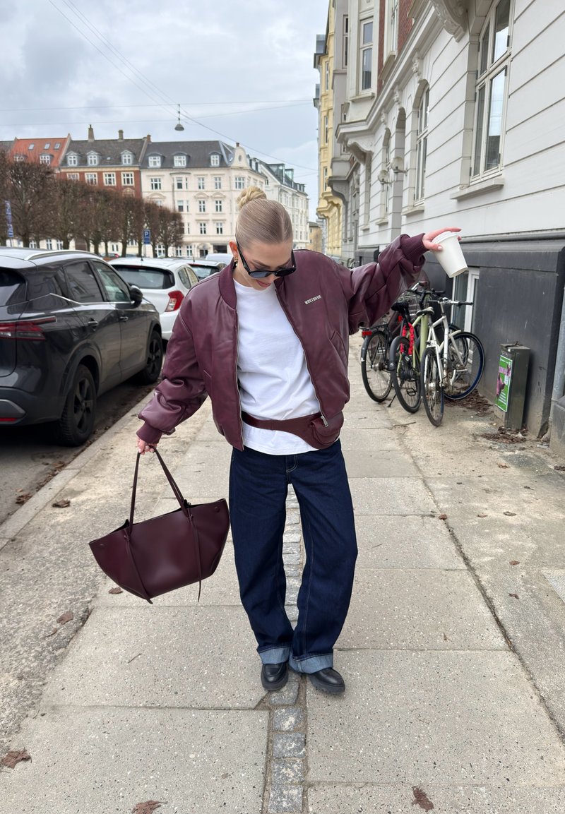 Jeune personne portant des lunettes de soleil et une veste bordeaux tient un sac bordeaux et une tasse blanche, debout sur un trottoir en ville à proximité de voitures et de vélos garés.