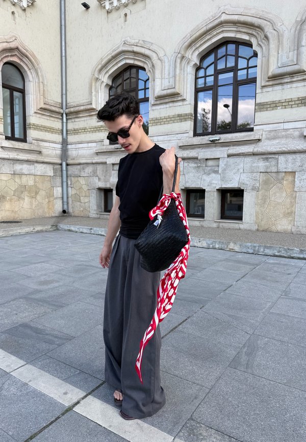 Une personne portant des lunettes de soleil, une chemise noire et un pantalon gris ample tient un sac noir tissé et une écharpe rouge et blanche devant un bâtiment en pierre.