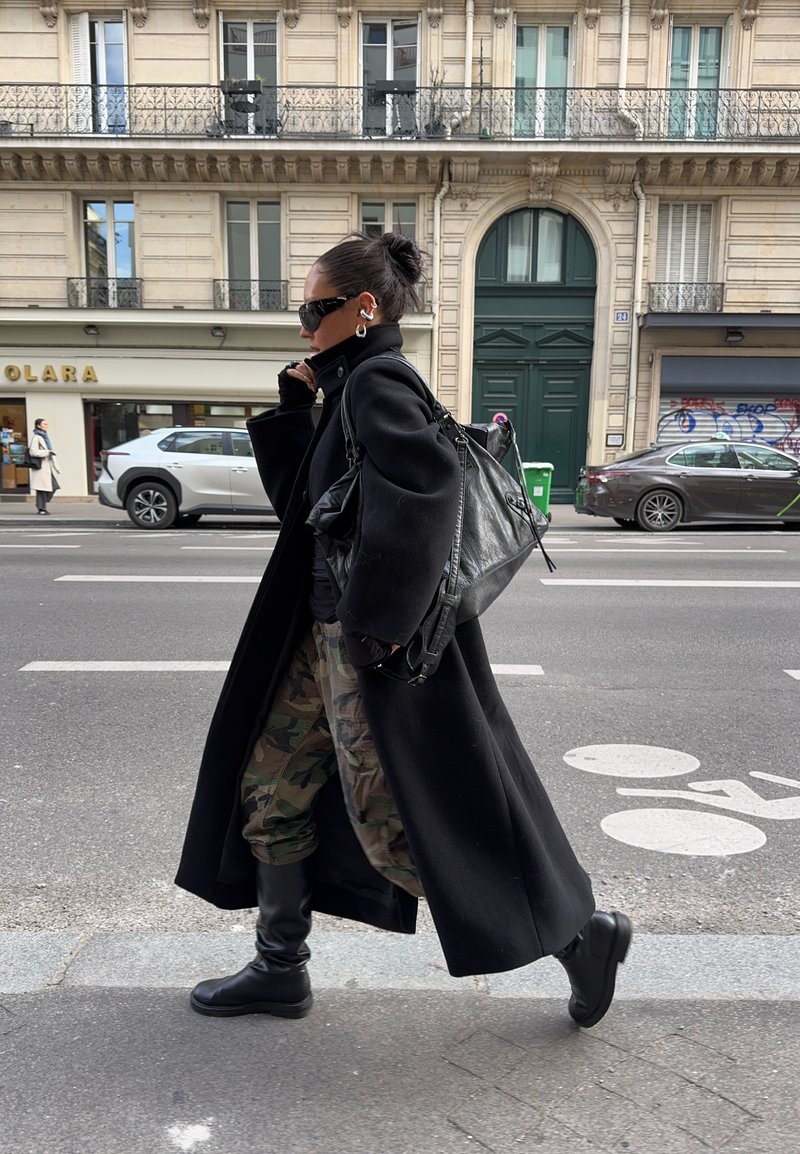 Femme en manteau noir, pantalon camouflage et bottes, marchant sur un trottoir en ville en portant un grand sac noir, avec des voitures et des bâtiments en arrière-plan.