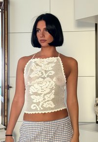 Ženska s kratkimi črnimi lasmi, oblečena v belo kvačkano halter-top in modre karirane hlače, stoji v notranjosti blizu okna.