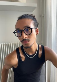 Lunettes rondes noires avec des verres teintés bleus, débardeur noir sans manches, colliers en perles et chaînes en argent superposés, teint de peau chaud, lumière naturelle.