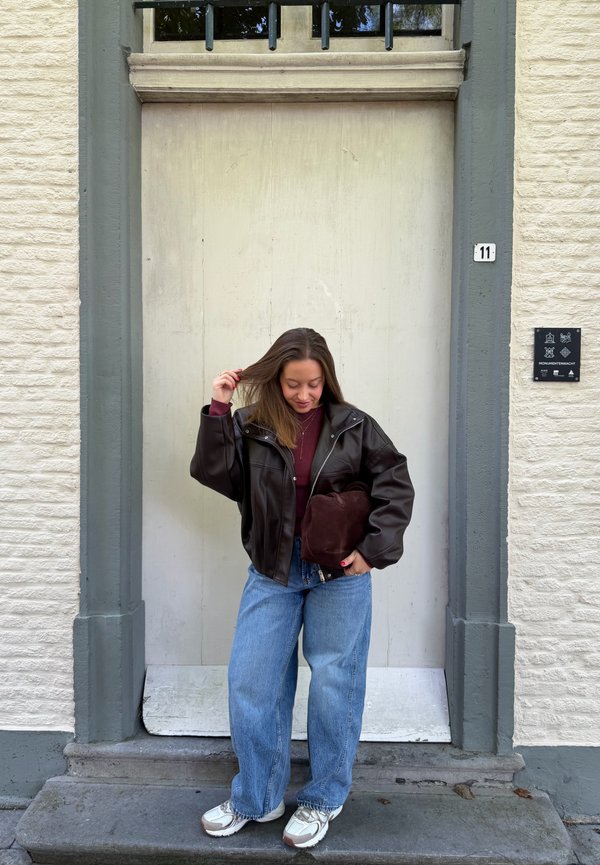Mujer de pie en la puerta vestida con una chaqueta de cuero oscura, suéter burdeos, vaqueros azules, zapatillas blancas, sosteniendo una bolsa marrón y tocándose el cabello.