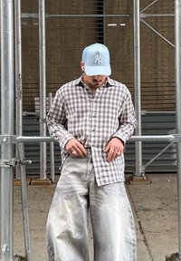 Kockás szürke és bézs gombos ing, bő, kifakult szürke farmernadrág, valamint világoskék baseballsapkás logóval. Ékszerek az ujjakon.