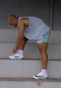 Idrettsutøver iført en lett grå tanktop og lysegrønne shorts, som knyter hvite sportssko med marineblå detaljer mens han står på betongtrapper.