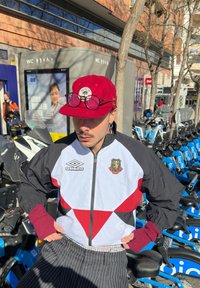 Boné vermelho com logotipo, casaco corta-vento preto e branco com padrões triangulares vermelhos e calças listradas. Os acess�órios incluem óculos de sol e luvas sem dedos.