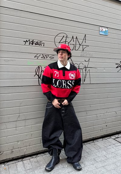 Joven con camiseta de rugby a rayas rojas y negras, vaqueros oscuros holgados, botas negras y gorra roja de béisbol, de pie frente a una pared cubierta de graffiti.
