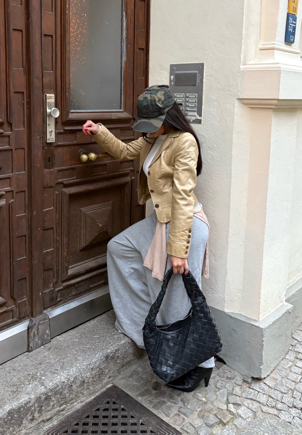 Woman in a beige leather jacket and grey trousers, holding a woven black handbag, near a wooden door with a large brass handle.