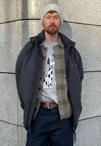 Doudoune noire portée sur une chemise à carreaux verte, t-shirt gris avec un motif de chien, jean bleu et bonnet gris clair.