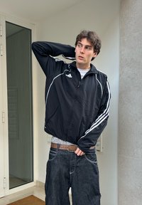 Veste de sport noire à zip avec des rayures blanches sur les bras, portée sur une chemise rayée et un jean en denim foncé ample, maintenu avec une ceinture marron.