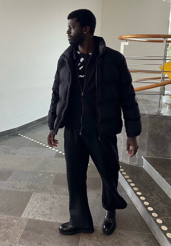 Uomo con giacca a piumino nera, pantaloni neri e scarpe nere lucide, in piedi all'interno vicino a una scala con corrimano in metallo e legno.