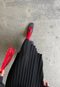 Falda negra con plisados, medias rojas, zapatos negros con correas decorativas y una bolsa roja con cremallera, sobre un suelo de hormigón.
