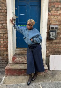 Denim bomberjakke med en afslappet pasform, kombineret med vidbenede mørke jeans. Sorte spidse sko, stående foran en blå dør med et nødudgangsskilt.