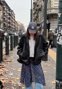 Black leather jacket with wide lapels, grey cap with logo, grey jumper, and a blue checked skirt over light blue jeans on a street with fallen leaves.