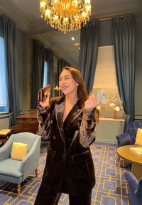 Žena v hnědém sametovém obleku se usmívá a drží peněženku, stojí v dekorovaném pokoji s lustr, modrými závěsy a čalouněnými židlemi.