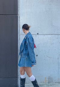 Jeans jakna z pasom, stilizirana kot kratka obleka. Vključuje rdečo torbico, črne čevlje do kolen in bele nogavice. Siva betonska ozadje.