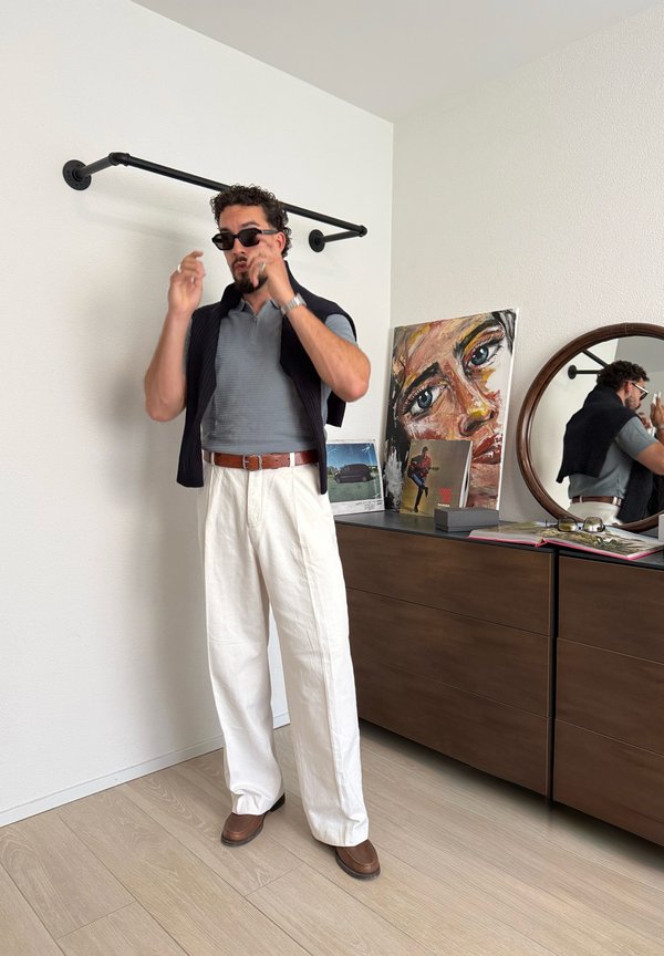 Mann mit Sonnenbrille, grauem Hemd, weißer Hose und braunen Schuhen richtet die Sonnenbrille in einem Zimmer mit Kommode, rundem Spiegel und farbigem Gemälde.