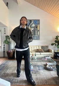 Brun læderjakke over en sort sweater, hvid skjorte og sorte bukser. Mørke sko. Moderne opholdsrum med et glas sofabord og planter.