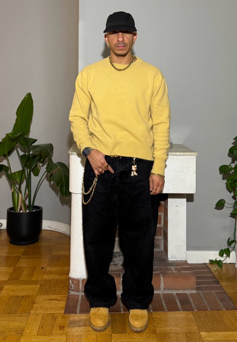 Maglione giallo, jeans neri larghi, stivali color cuoio, cappello nero, collana a catena dorata e dettaglio portachiavi sui jeans. Ambientazione interna con piante.