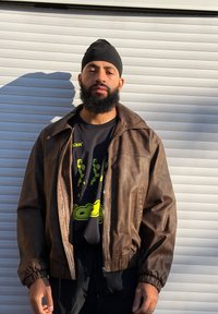 Homme barbu portant un turban noir, une veste en cuir marron et un t-shirt graphique noir, debout devant une porte de garage en métal blanc sous la lumière du soleil.