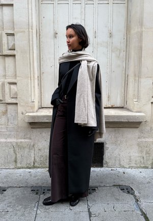 Mujer con abrigo largo negro, pantalones oscuros, guantes negros y bufanda crema sobre los hombros, de pie frente a un edificio de piedra clara con persianas cerradas.