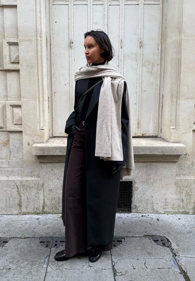 Mujer con abrigo largo negro, pantalones oscuros, guantes negros y bufanda crema sobre los hombros, de pie frente a un edificio de piedra clara con persianas cerradas.
