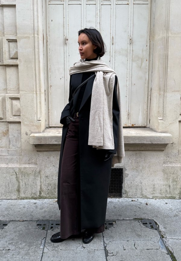 Mujer con abrigo largo negro, pantalones oscuros, guantes negros y bufanda crema sobre los hombros, de pie frente a un edificio de piedra clara con persianas cerradas.