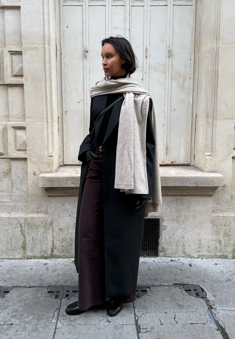 Femme portant un long manteau noir, un pantalon foncé, des gants noirs, une écharpe crème posée sur les épaules, debout contre un bâtiment en pierre claire avec des volets fermés.