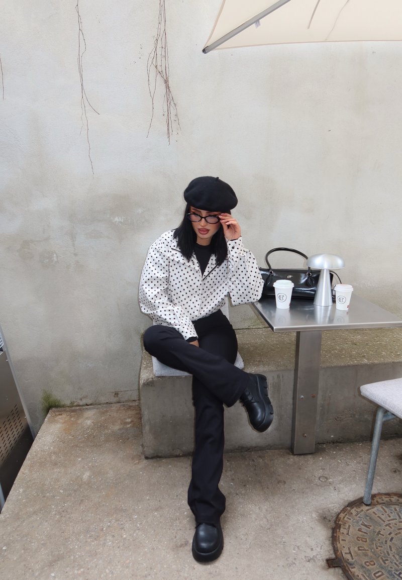 Femme en béret noir et veste à pois assise en tailleur sur un banc en béton à côté d'une table avec deux tasses de café et un sac à main.