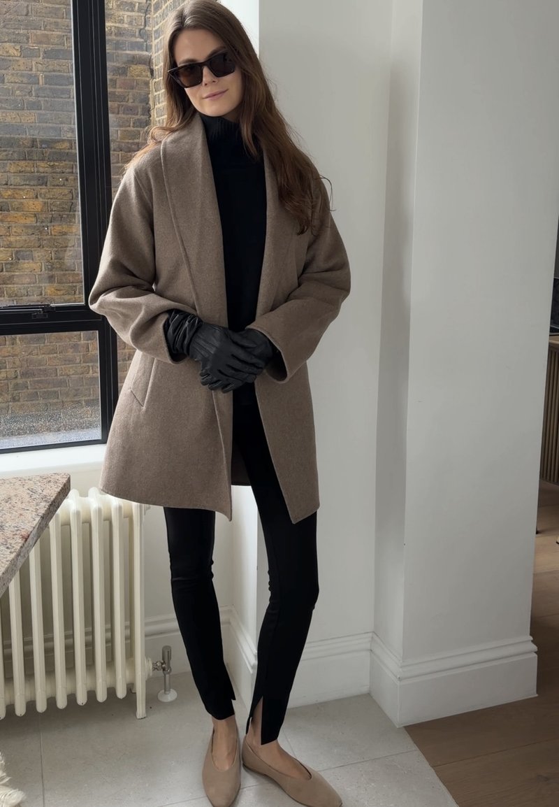 Femme portant un manteau beige oversize, un col roulé noir, des gants noirs, un pantalon noir slim avec des fentes aux chevilles, des ballerines beiges et des lunettes de soleil foncées à l'intérieur.