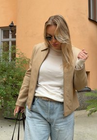 Beige nahkatakki, jossa on vetskari edessä, valkoisen paidan päällä. Vaaleansiniset farkut ja mustat aurinkolasit. Taustalla näkyy vihreyttä ja seiniä.
