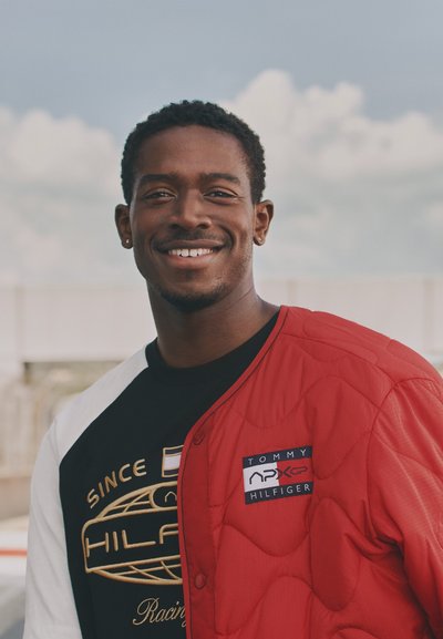 Chaqueta acolchada roja con el logotipo de "Tommy Hilfiger" y camisa en blanco y negro con bordados dorados, en un fondo de cielo nublado.