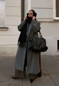 Grijze lange jas, zwarte franjesjaal, zwarte oorwarmers, op maat gemaakte broek, puntige zwarte hakken en een zwarte handtas met studs. Soepele stoftexturen.