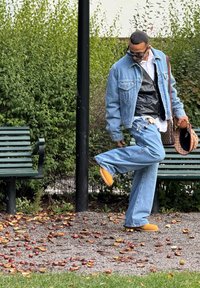 Denimjacke über einer schwarzen Weste, lockere blaue Jeans und gelbe Schuhe. Steht neben grünen Bänken, umgeben von gefallenem Laub.