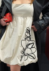 Femme portant une robe blanche brodée de fleurs noires, une veste en cuir noire, tenant un sac à main rouge avec une bandoulière chaîne dorée.