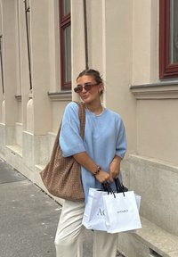 Maglione oversized blu chiaro, borsa tote beige in tessuto, borse della spesa bianche con manici neri e pantaloni color crema. Dettagli di texture sottili.