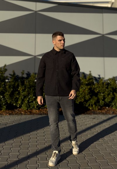 Chaqueta negra abotonada con dos bolsillos en el pecho; jeans gris; zapatillas blancas; de pie sobre un pavimento de patrones con un fondo geométrico.