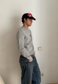 Šedá mikina s žebrovanými manžetami a tmavě modré džíny s širokými nohavicemi. Černá čepice s červeno-bílým motivem vlajky Spojeného království. Stojí opřený o zeď.