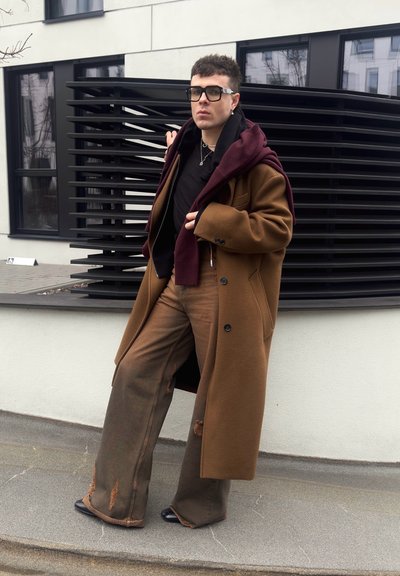 Persona con abrigo marrón holgado, pantalones anchos rasgados, camisa oscura, suéter burdeos sobre los hombros, gafas negras grandes, de pie al aire libre.