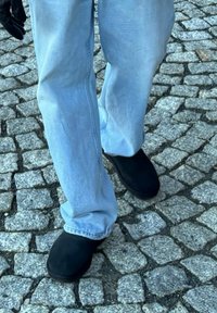 Črne natikače brez pet, kombinirane z svetlo modrimi kavbojkami, ki imajo odrezane robove, stojijo na strukturirani kamniti površini.