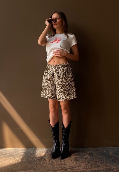 Camiseta blanca corta con gráfico de bota roja, combinada con shorts de leopardo en color beige. Las botas negras hasta la rodilla con tachuelas plateadas completan el conjunto.