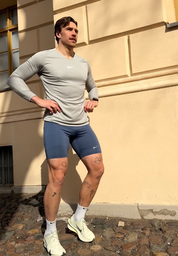 Atletische man met tatoeages staat zelfverzekerd buiten, handen in de zij, gekleed in een grijs shirt, blauwe shorts, witte sneakers en witte sokken.