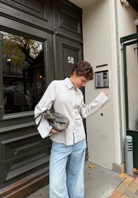 Csíkos, világos bézs hosszú ujjú gombos ing, világoskék, bőszárú farmerral párosítva. Egy kis, fémes szürke kézitáskát tart a kezében.